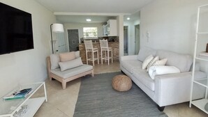 A modern living room with a sofa, a coffee table, and a TV. There is a kitchen area in the background with a dining set. - Idyllic serene secure condo minutes from beach & amenities.  (Bridgetown)