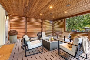 Um deck de madeira com área de estar ao ar livre, lareira e vista para árvores.