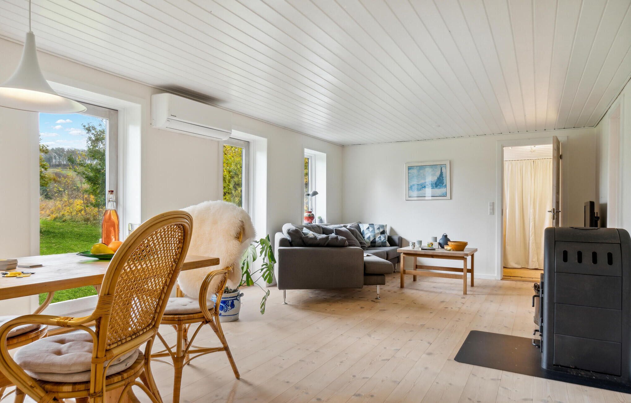 Een ruime woonkamer met een eettafel, rieten stoelen, een grijze bank en een zwarte grillplaats.