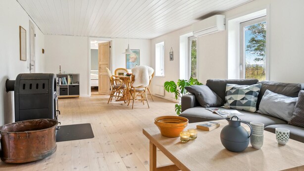 Un amplio salón con piso de madera, un sofá, una mesa con adornos y vistas al exterior a través de grandes ventanales.