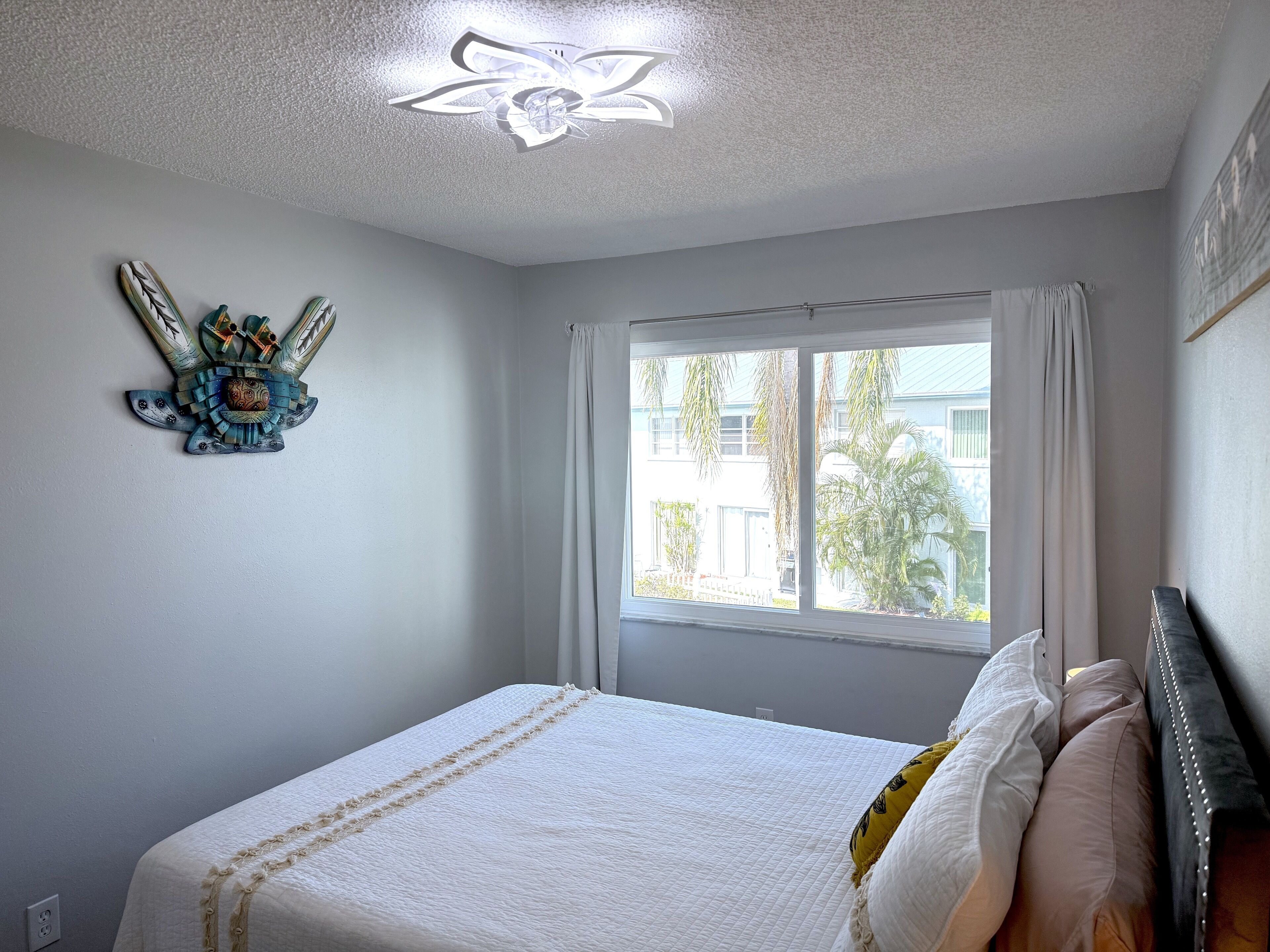 A bedroom with a bed, a ceiling fan, a window with curtains, and a wall decoration.