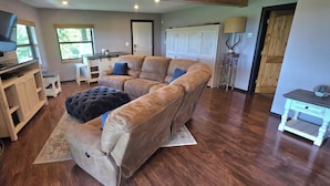 A living room with a large leather sofa, a flat-screen TV, wooden flooring, a kitchen area, and a dining table. - Lakeview Hideaway - Amazing View of Lake of the Arbuckles (Sulphur)