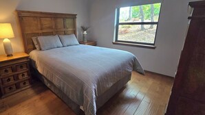 A bedroom with a wooden headboard, a bed with a grey bedspread, a nightstand with a lamp, and a window with a view of trees. - Lakeview Hideaway - Amazing View of Lake of the Arbuckles (Sulphur)