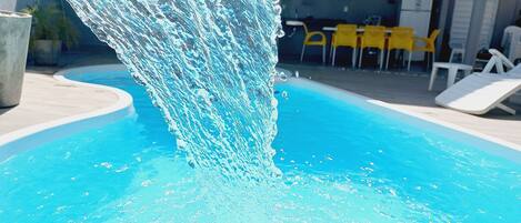 A swimming pool with a water feature, surrounded by lounge chairs and yellow tables.
