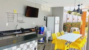 A compact kitchen with a refrigerator, sink, and a flat-screen TV. There is a dining area with yellow chairs and a table. - Casa Paraíso das Conchas (Barra de São Miguel)