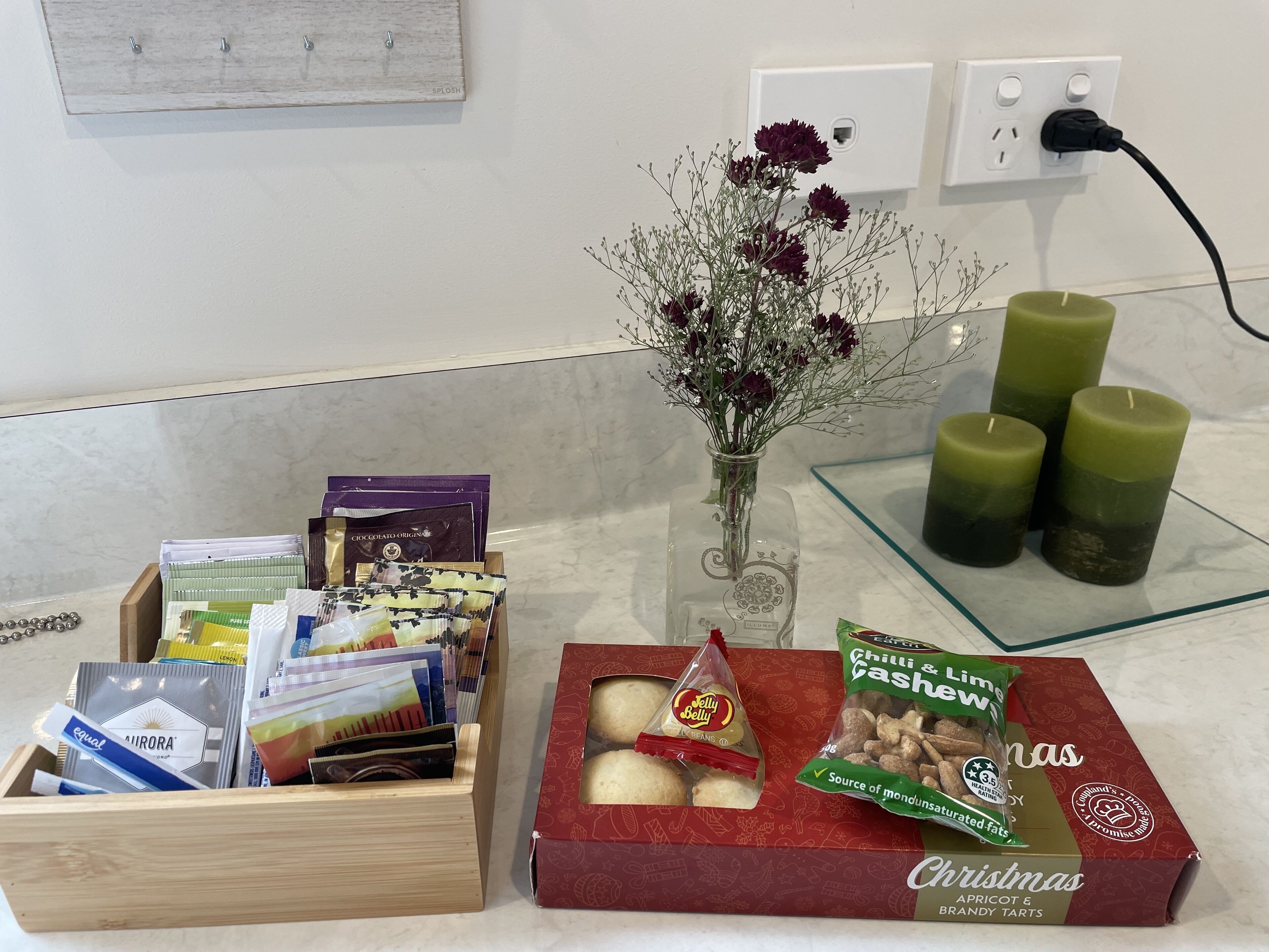 A wooden box with assorted items, a red box with chocolates, a vase with flowers, and three green candles on a table.