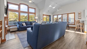 A spacious living room with a large blue sofa, a dining area with wooden chairs, and large windows offering views of trees. - 7 Ensuites + Private Pool + 5 Min Walk to Downtown (Kitty Hawk)