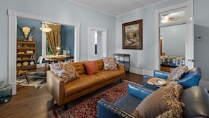 Een woonkamer met een bruine bank, blauwe fauteuils en een rood geweven vloerkleed. Je ziet een houten boekenkast en een kleine tafel met een lamp.