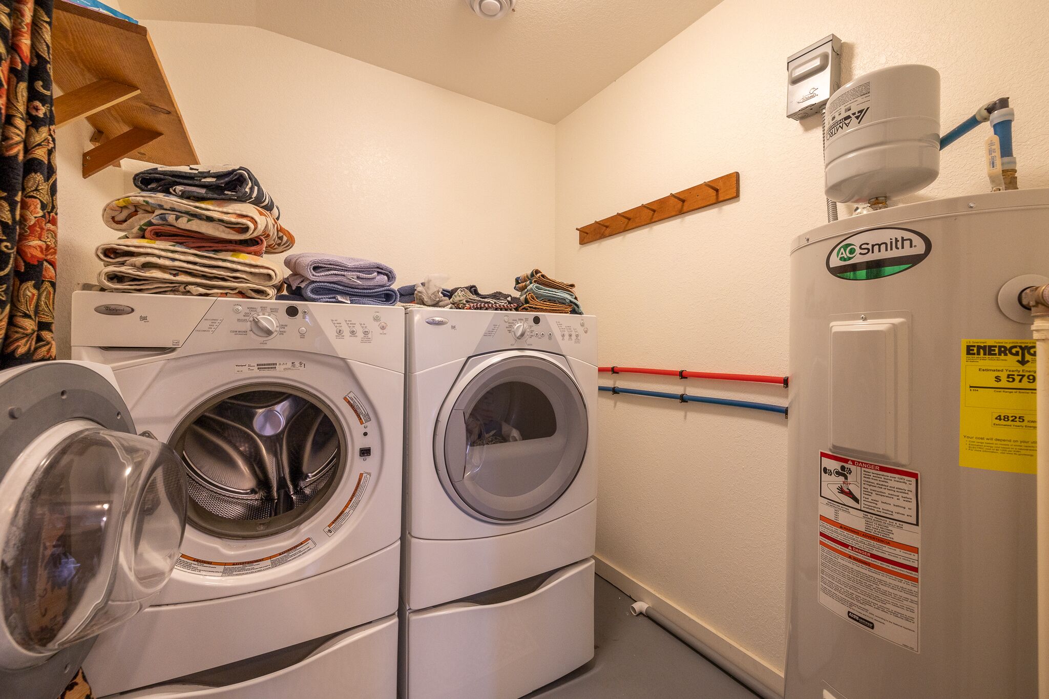A laundry area with two stacked front-loading washers and dryers, a water heater, and a wooden shelf with folded clothes.