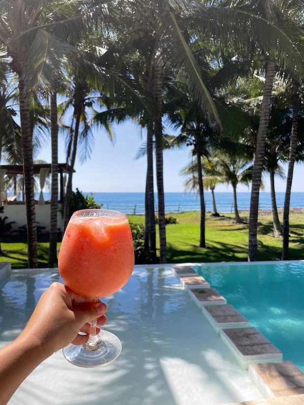 Una mano sostiene un vaso con una bebida rosa, con vista a una piscina, palmeras y el océano.