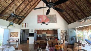 Uma sala de estar ampla com teto alto de palha, uma mesa de jantar central e cozinha ao fundo.