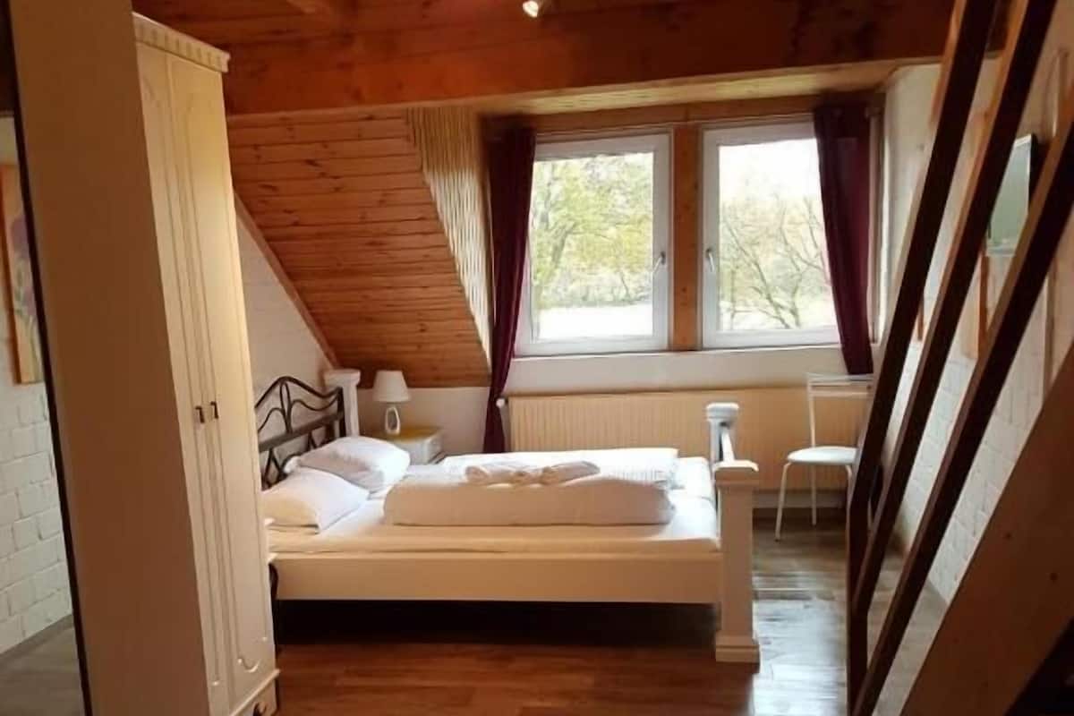 Quarto com teto de madeira, cama, janela com cortinas e radiador.