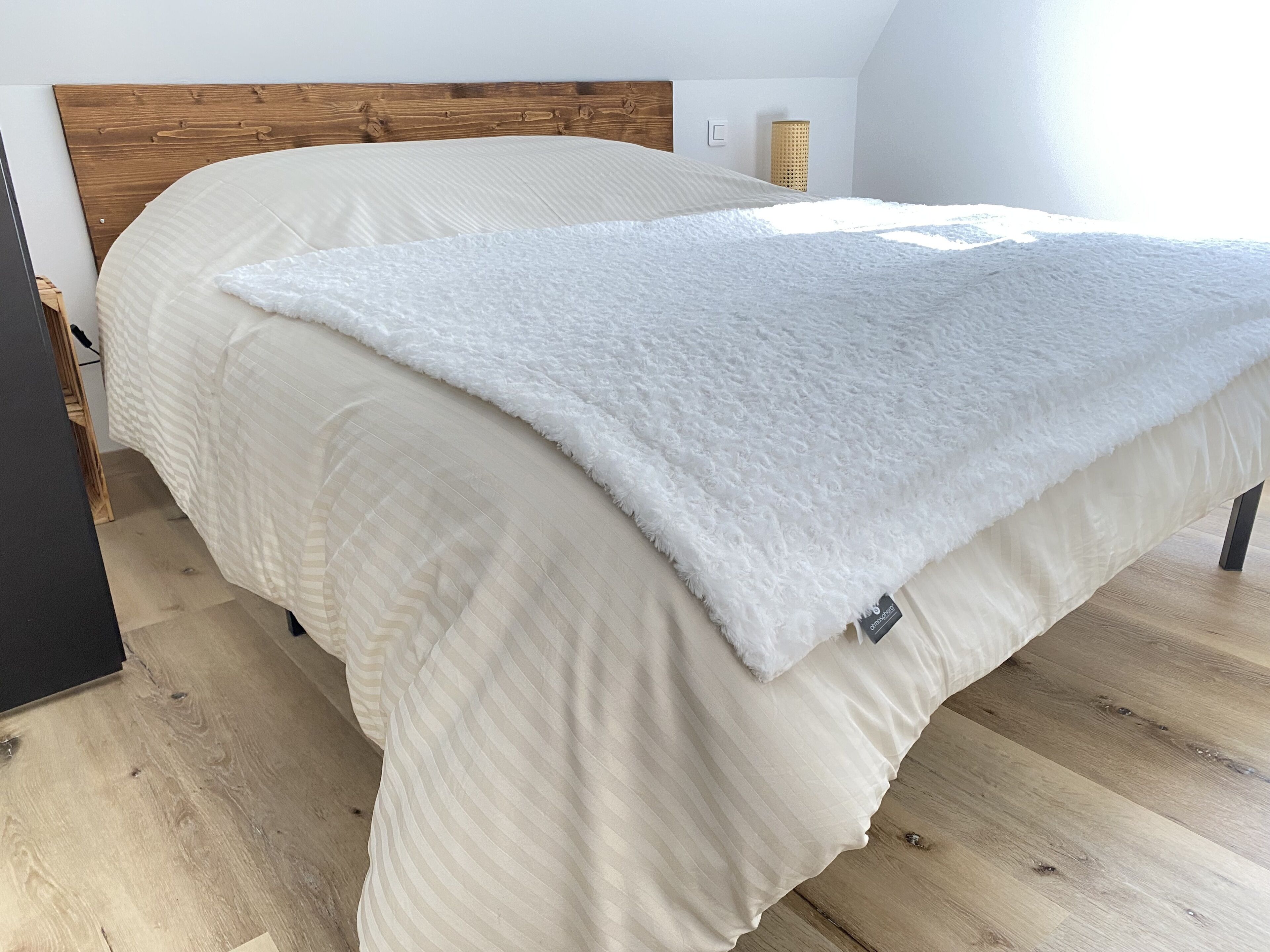 Un lit bien fait, avec une couette blanche et une couverture blanche moelleuse, une tête de lit en bois et un plancher en bois de couleur claire.