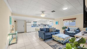 Ein geräumiges Wohnzimmer mit einem blauen Sofa, passendem Hocker und einem Essbereich mit weißem Tisch und Stühlen. Im Hintergrund ist eine Küche zu sehen.