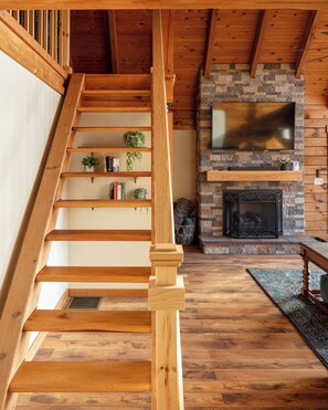 A wooden staircase with a built-in bookshelf, a stone fireplace, and a television mounted above it. - New Hot Tub - Sweet Pines Cabin - Sleeps up to 8 (Patriot)