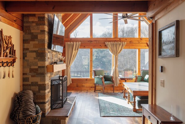 A cozy cabin interior with a stone fireplace, wooden beams, a dining area, and large windows. - New Hot Tub - Sweet Pines Cabin - Sleeps up to 8 (Patriot)