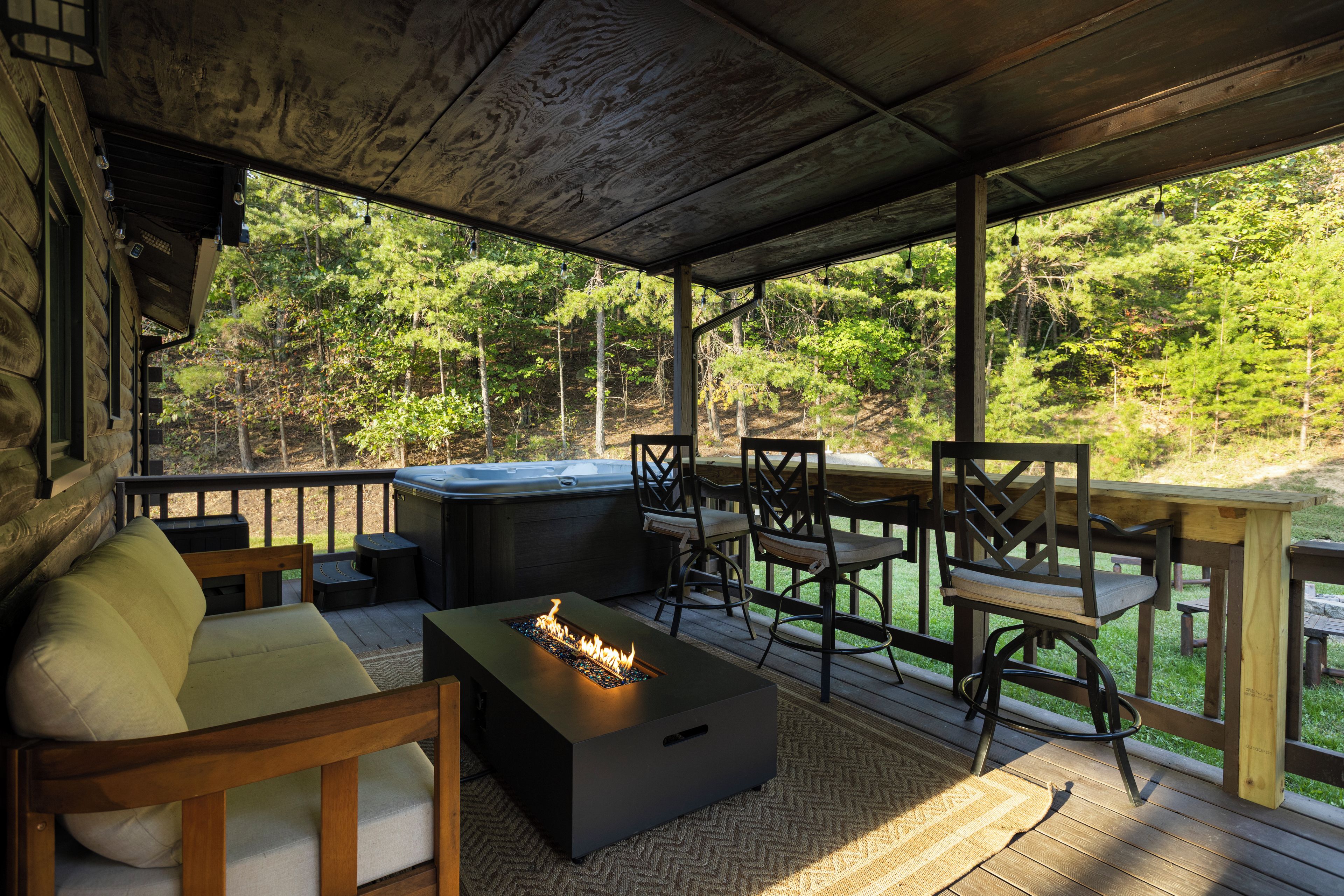 Un espace extérieur aménagé, partiellement abrité, avec un foyer, un jacuzzi et une vue sur une zone boisée.