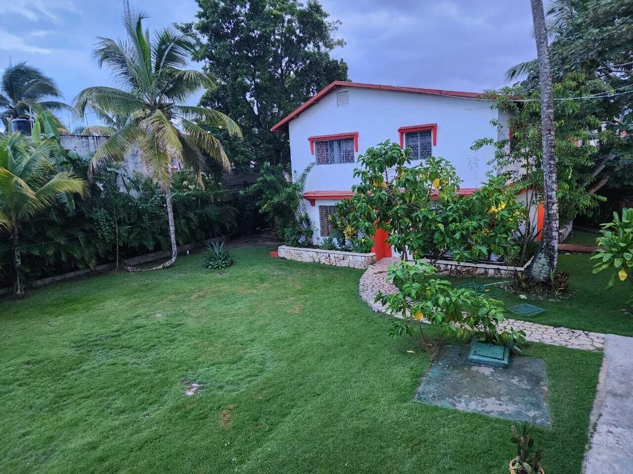 Un bâtiment blanc orné de bordures rouges et doté d’une porte rouge, entouré d’une pelouse bien entretenue et d’une végétation tropicale.