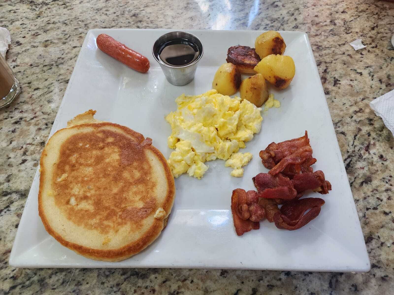 A breakfast plate with pancakes, scrambled eggs, bacon, sausages, and roasted potatoes.