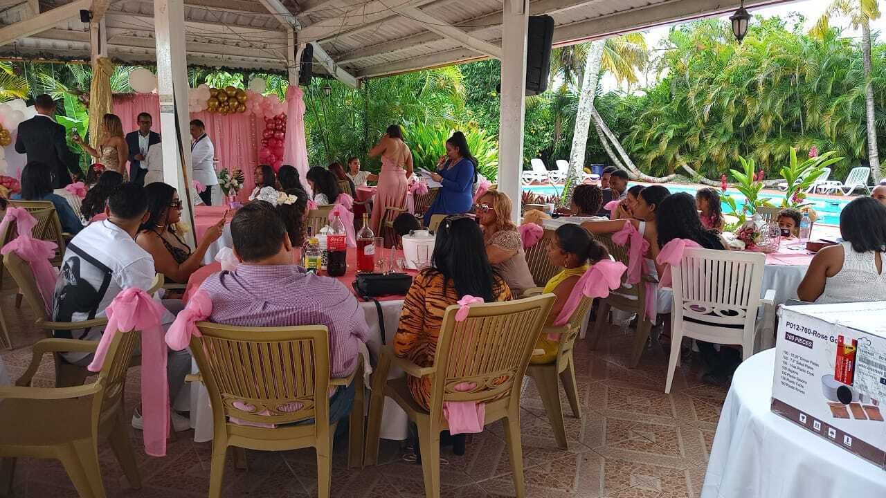 Evento al aire libre con personas sentadas en mesas, algunas con manteles blancos, y sillas adornadas con cintas rosas.