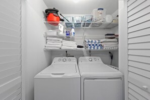 Un cuarto de lavandería con dos lavadoras blancas, repisas superiores con toallas, una botella de detergente y una aspiradora roja.