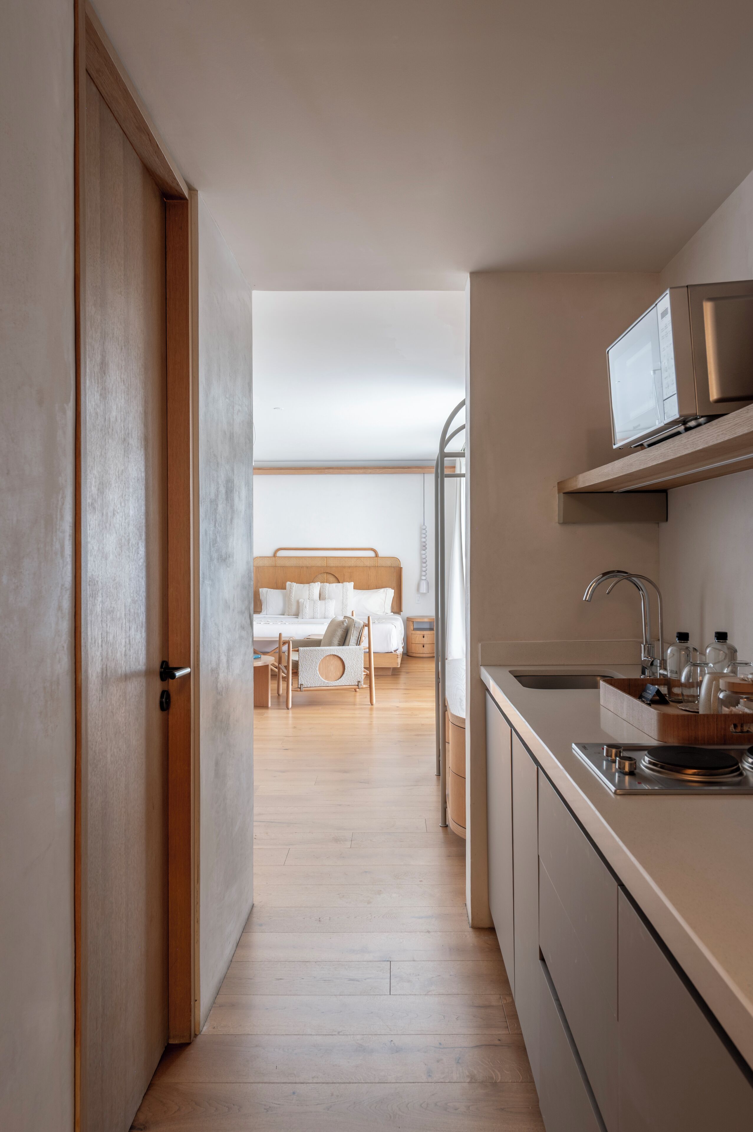 A modern hotel room with a wooden floor, a bed with white linens, a small seating area, and a kitchen with a sink and microwave.