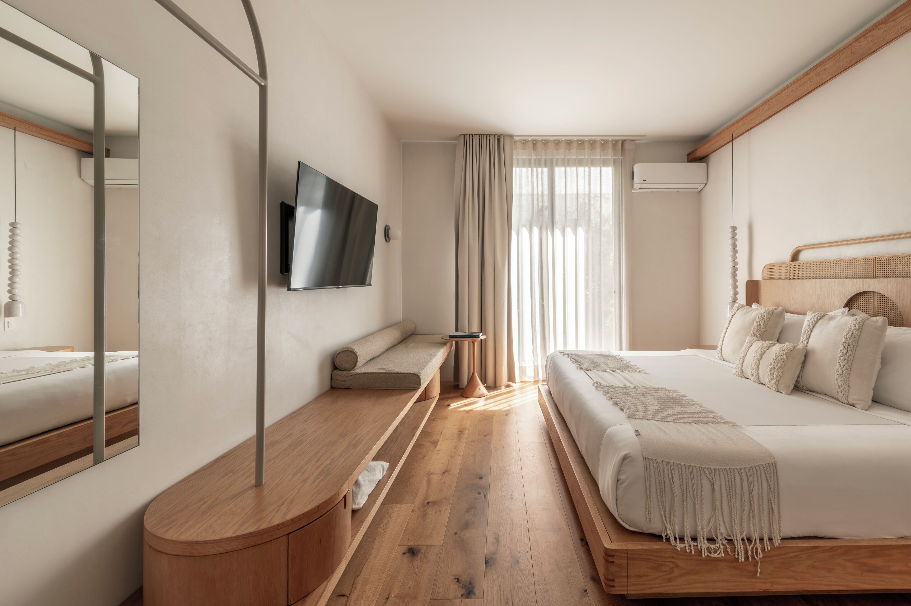 A modern bedroom with a wooden floor, a large bed with white bedding, a TV mounted on the wall, and a bedside table with a lamp.