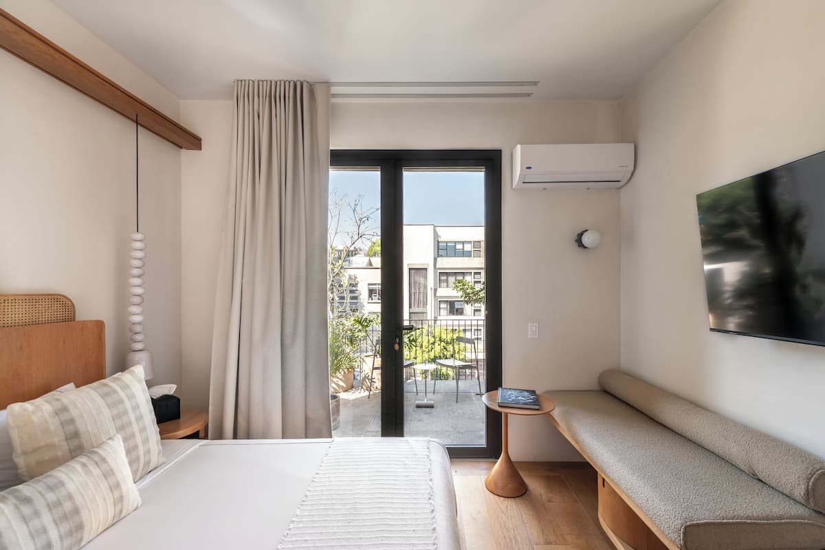 Une chambre d’hôtel moderne équipée d’un lit, d’une banquette, d’une télévision et d’un balcon offrant une vue sur les bâtiments.