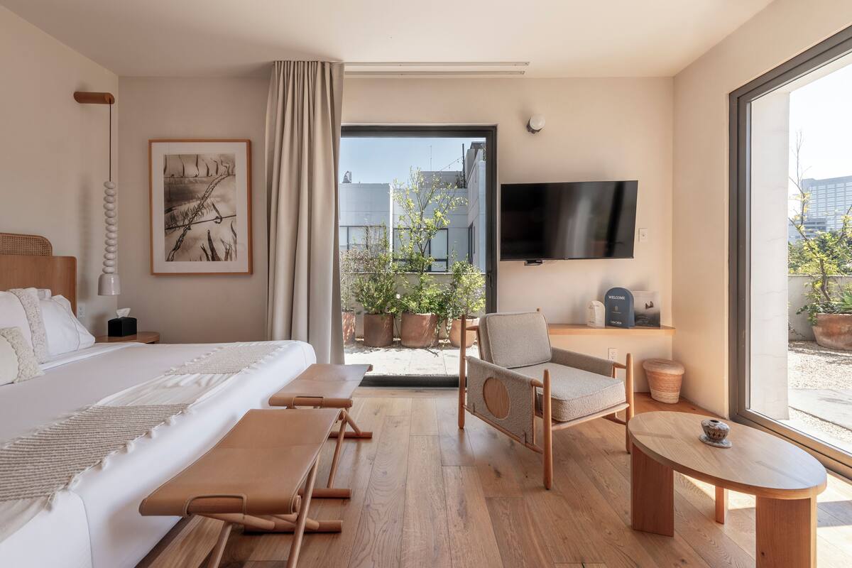 Moderni hotellihuone, jossa on suuri sänky, oleskelualue pöydällä, seinälle asennettu televisio ja näkymä ulos.