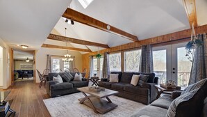 A spacious living room with a high ceiling, wooden beams, large windows, and a cozy seating area. - Welcome to our Pocono Cottage (Tobyhanna)
