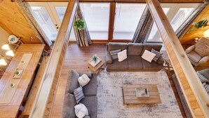 A loft-style room with a wooden ceiling, large windows, a gray sectional sofa, a coffee table, and a dining table with chairs. - Welcome to our Pocono Cottage (Tobyhanna)