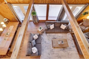 A loft-style room with a wooden ceiling, large windows, a gray sectional sofa, a coffee table, and a dining table with chairs. - Welcome to our Pocono Cottage (Tobyhanna)