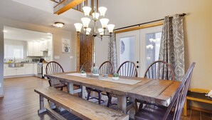 A spacious dining area with a wooden table and benches, a chandelier, and a view of the kitchen in the background. - Welcome to our Pocono Cottage (Tobyhanna)