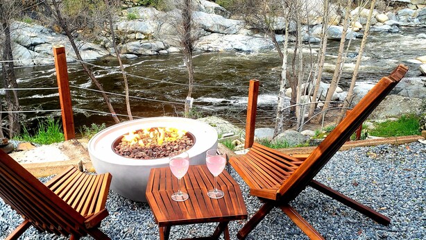 A fire pit with two wooden chairs and a table, surrounded by a gravel area with a view of a river and rocks. - Retro River House! Waterfront, Firepit, BBQ, Gameroom, 1 min to Sequoia Park.  (Three Rivers)