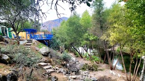A natural outdoor area with a wooden deck, stone steps, and a river. - Retro River House! Waterfront, Firepit, BBQ, Gameroom, 1 min to Sequoia Park.  (Three Rivers)