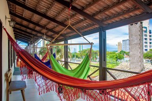 A hammock in a covered outdoor area with a view of tall buildings and palm trees. - Pousada Flat Castor (Natal)