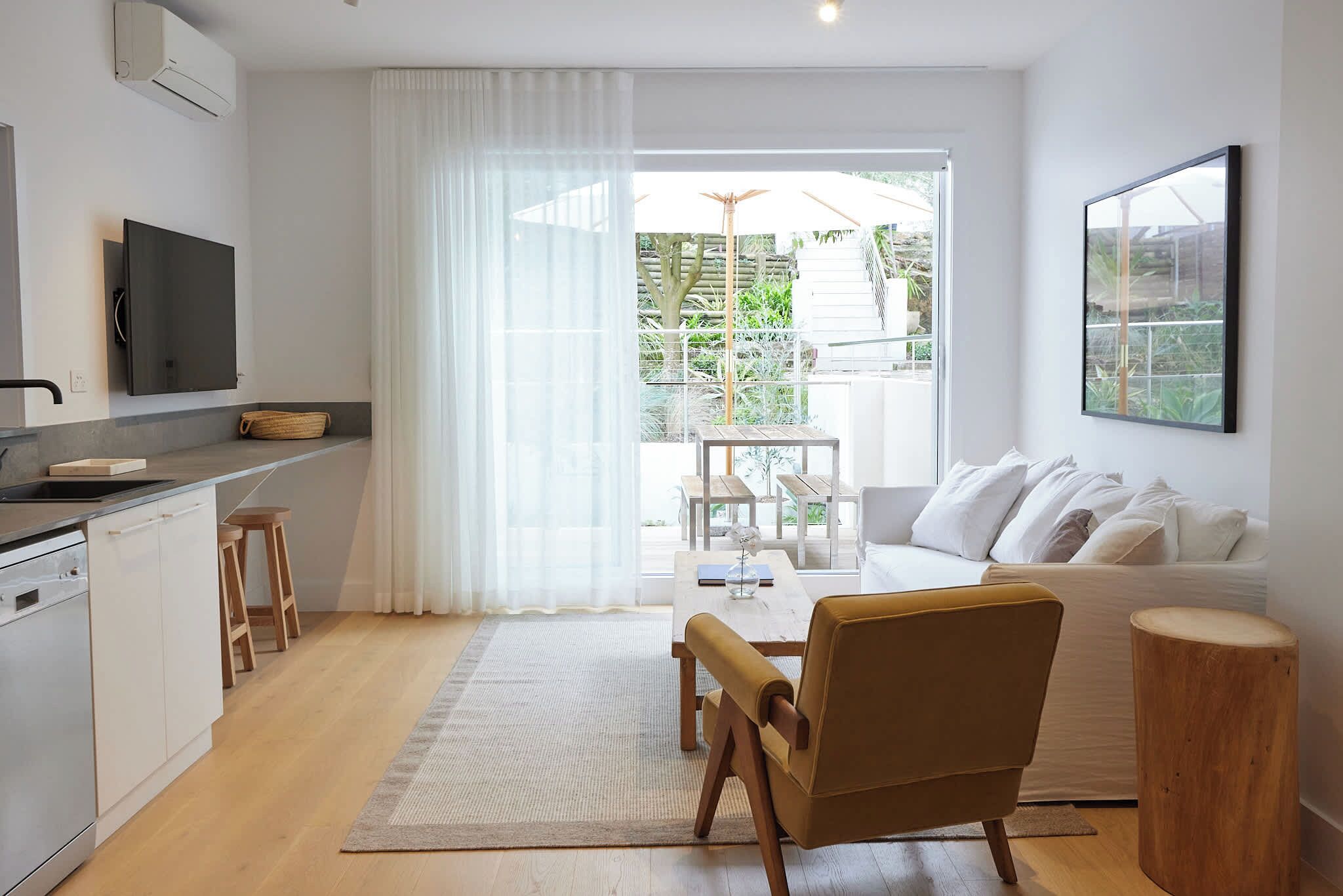 A modern kitchen with a flat-screen TV, wooden stools, and a dining area with a sofa and coffee table.