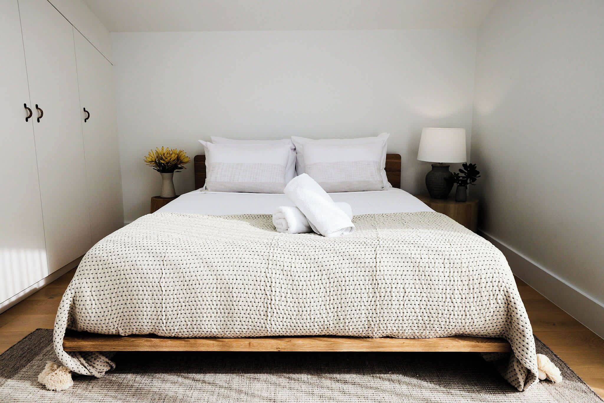 Una cama bien hecha con edredón blanco y almohadas, una lámpara de noche y un jarrón con flores sobre una mesita.