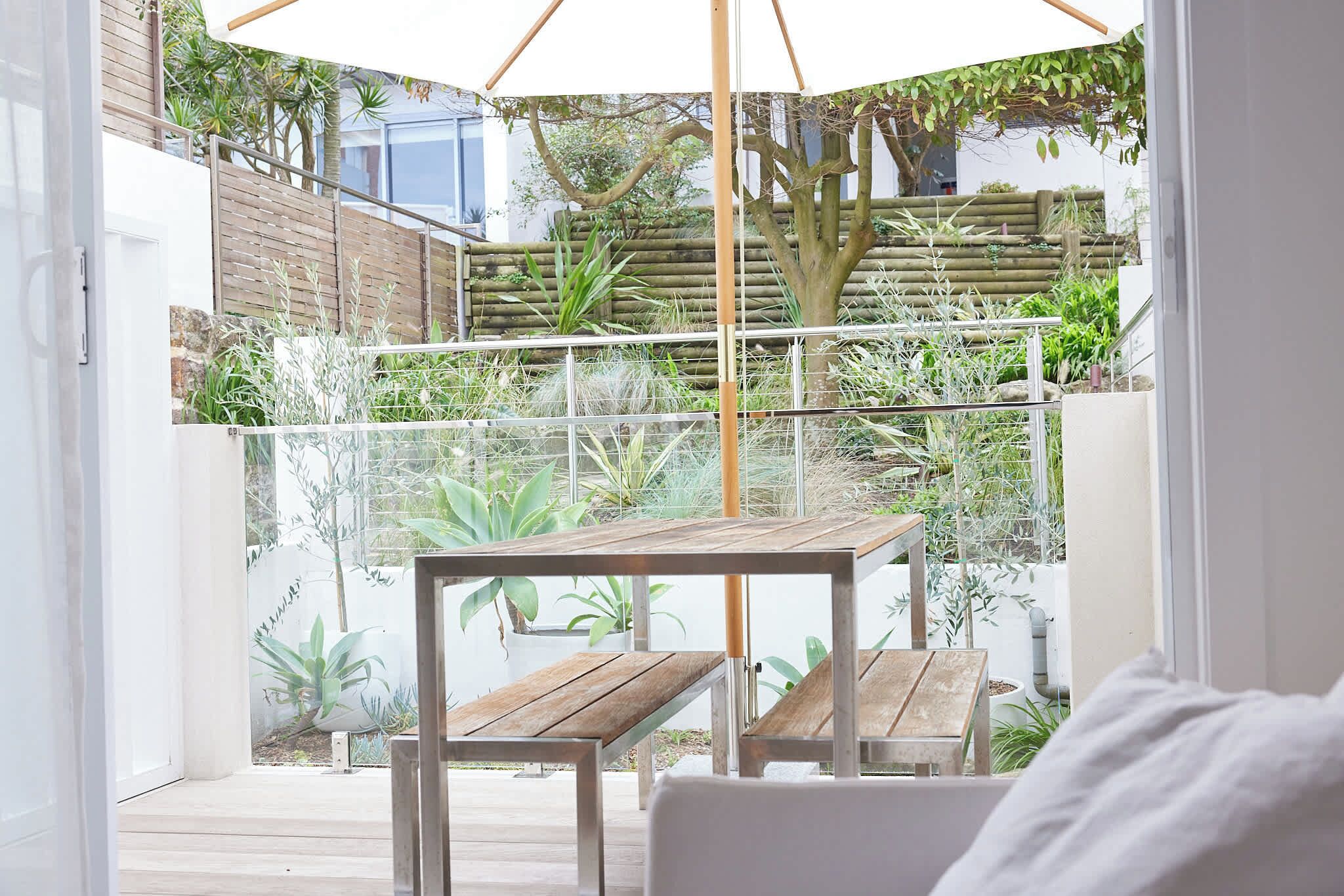 Un patio exterior moderno con una mesa y sillas de madera, rodeado de diversas plantas y una cerca de bambú.