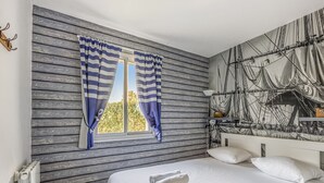 Una camera da letto con un letto, biancheria bianca, una finestra con tende a righe, un muro decorato con un murale a tema nautico e un termosifone.