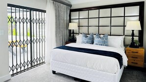 A hotel room with a large bed, two bedside tables with lamps, a headboard with a grid pattern, a view through a barred door, and a small wooden nightstand. - Staycation With Back Up Power (Germiston)
