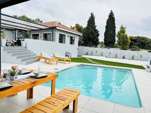 A modern pool area with a wooden deck, lounge chairs, and a dining table. - Staycation With Back Up Power (Germiston)