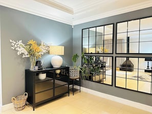 A modern living room with a black console table, a large mirror, and a view of a fireplace through a grid-patterned window. - Staycation With Back Up Power (Germiston)