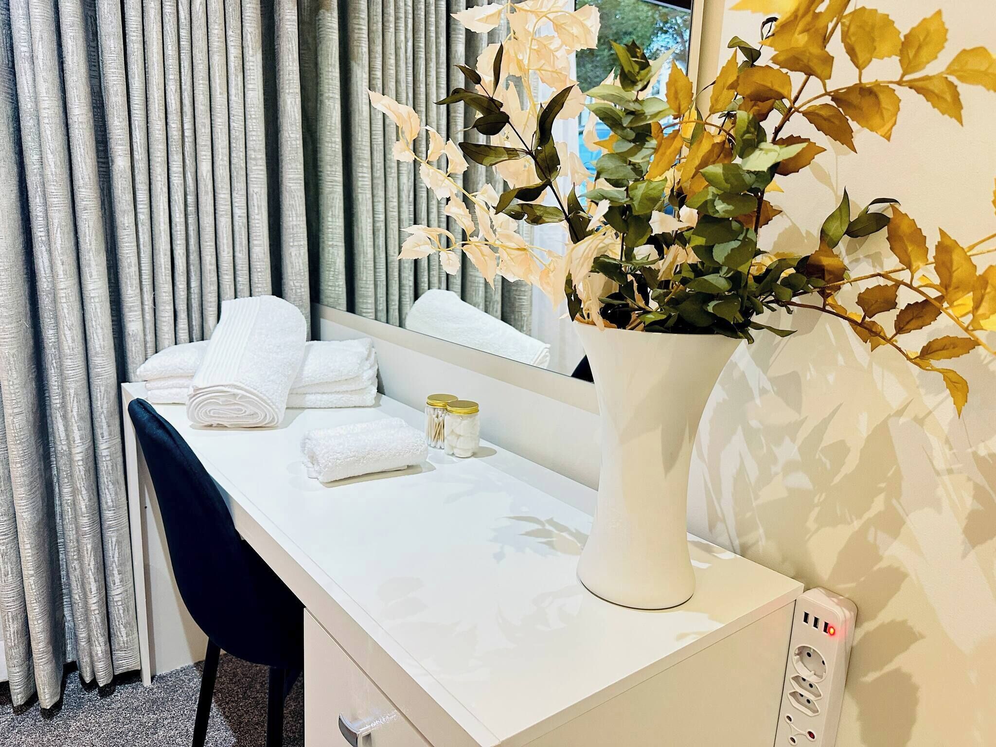 A hotel room with a desk, towels, a vase with flowers, a mirror, and a power outlet.