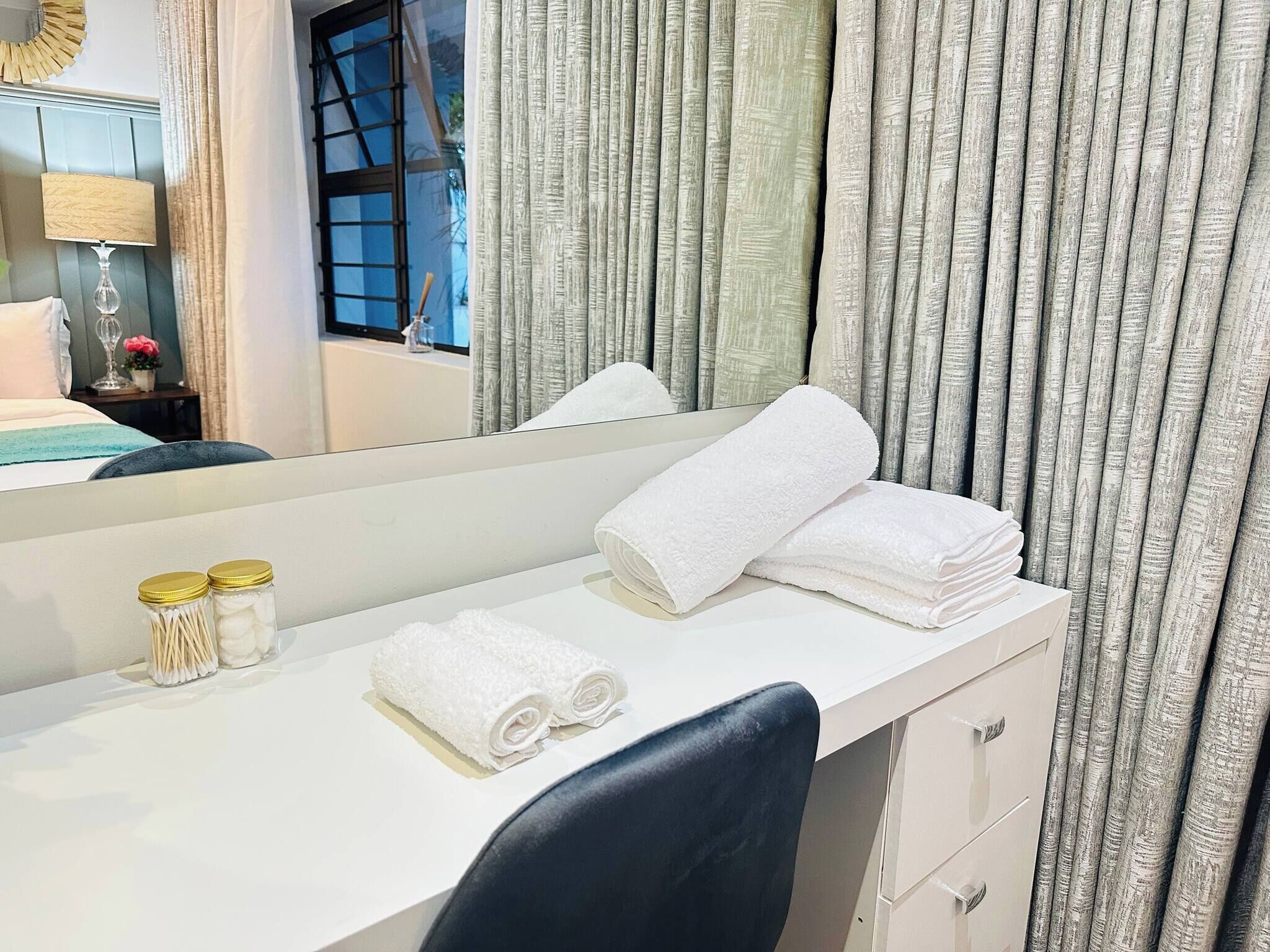 A hotel room with a white vanity, a mirror, a pair of rolled towels, and a window with curtains.