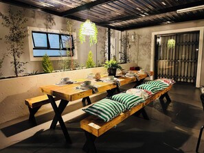 A dining area with a long wooden table, benches, potted plants, and hanging lights. - Staycation With Back Up Power (Germiston)