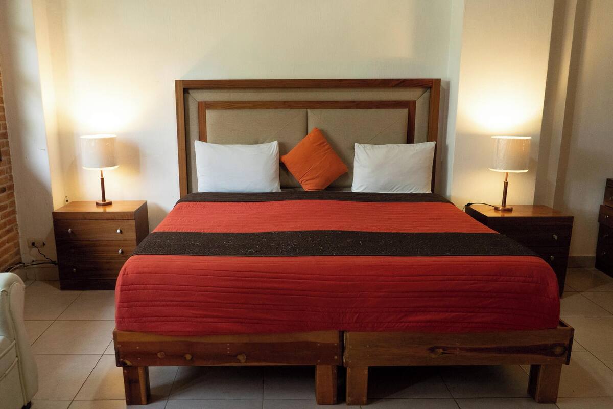 A neatly made bed with a wooden headboard, two white pillows, and an orange accent pillow. There are two bedside tables with lamps, a wooden bench at the foot of the bed, and a brick wall in the background.