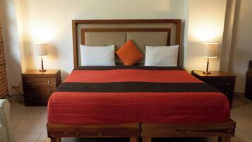 Una cama bien hecha con cabecera de madera, dos almohadas blancas y una almohada anaranjada decorativa. Hay dos mesitas de noche con lámparas, un banco de madera al pie de la cama y una pared de ladrillo al fondo.