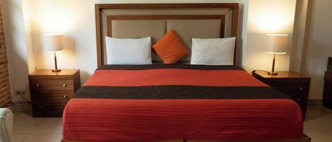 Cama bem arrumada com cabeceira de madeira, duas almofadas brancas e uma almofada laranja decorativa. Há duas mesas de cabeceira com abajures, um banco de madeira no rodapé da cama e uma parede de tijolos ao fundo.