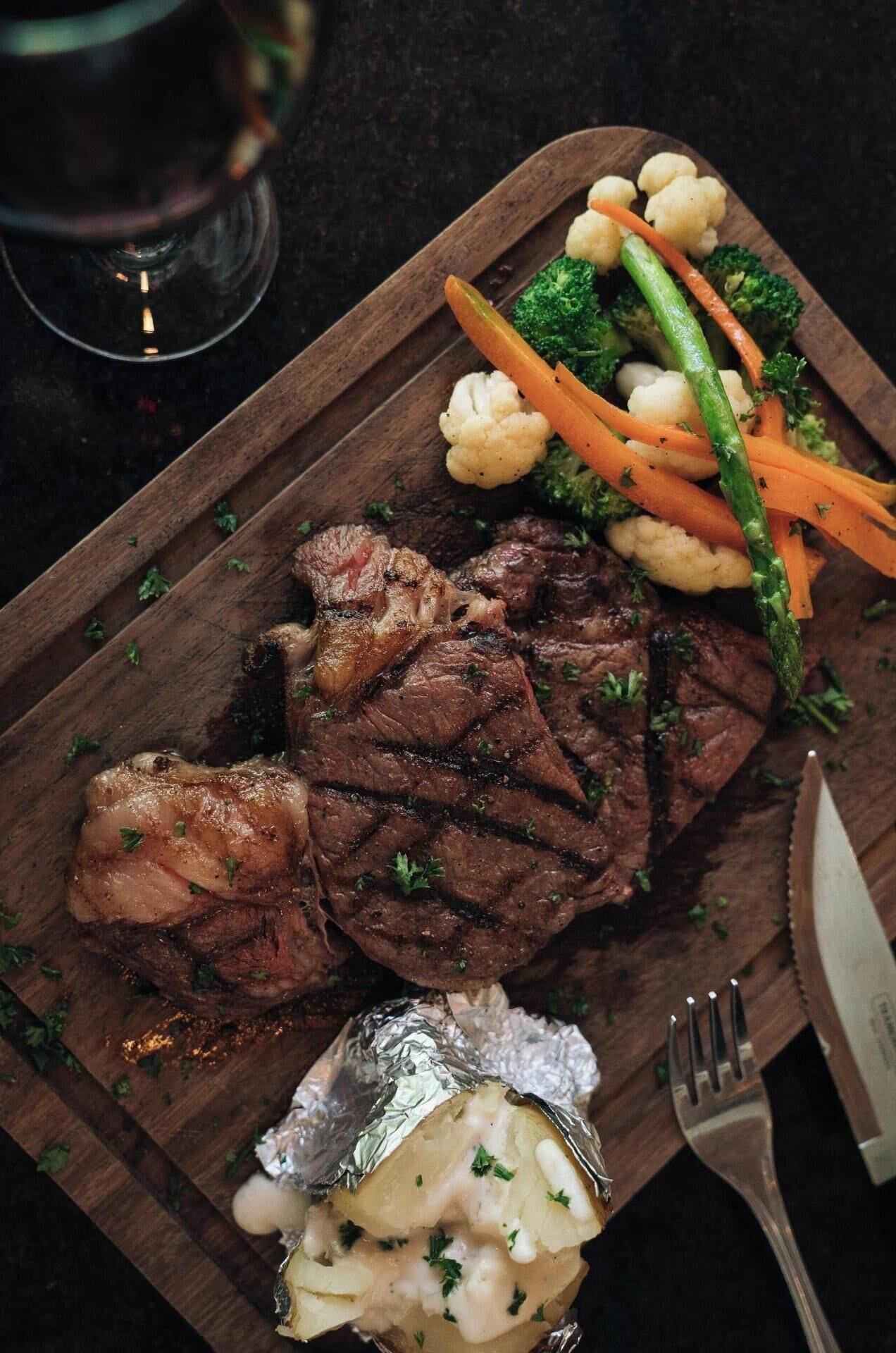 A wooden cutting board with a steak, vegetables, and foil-wrapped potatoes.